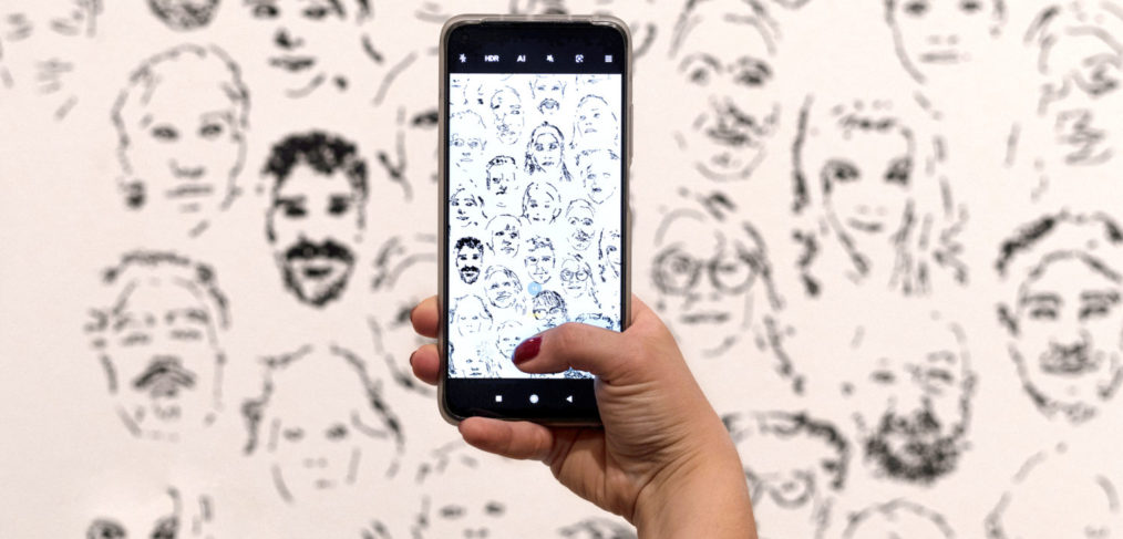 A phone is held up in front of a white wall with lots of faces drawn on it. The phones camera is enabled and so shows the faces in sharp focus.