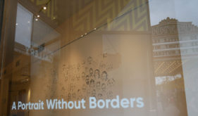Looking through a window at white walls with faces drawn in black lines. On the window are reflections of the ornate front entrance of The Flynn Theatre, the name of which you can see written in reverse in the reflection. On the window is a decal that reads "A Portrait Without Borders"