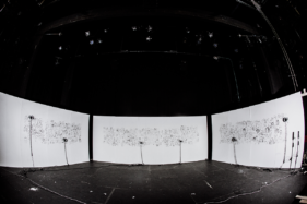 3 temporary white walls, each 6m wide and 2.4m high, stand on a large empty stage. On the walls are 500 faces drawn in black lines