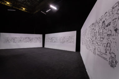 3 temporary white walls each 6m wide and 2.4m high stand on a large empty stage. On the walls are 500 faces drawn in black lines