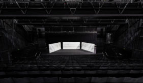 Three temporary white walls sit on a stage in a deserted auditorium with hundreds of empty seats facing the stage. Hundreds of faces are drawn on the walls.