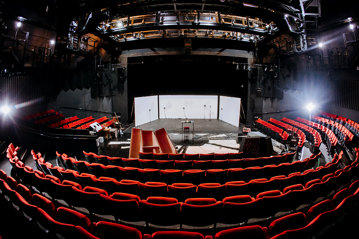 Wide-angled photo of a deserted auditorium. empty red seats face a stage with white walls where wall drawing machines draw portraits of the public