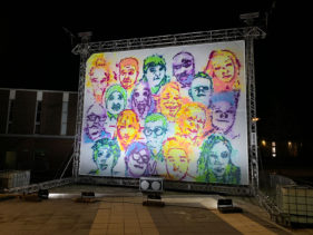 18 brightly coloured faces, spray-painted onto a 10m x 8m canvas are lit up. They glow in the darkness.
