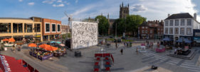 A 10m x 8m canvas of faces pulled taught across a truss structure, stands in the centre of a civic square, people eating at restaurants under umbrellas in the evening sun to the left and far right, a church and trees to the right of the canvas.