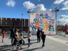 Outside, several people, parents and children, walking towards a 10m x 8m canvas on which are painted about 15 different faces in bright colours. Behind is blue sky and the sun is shining.
