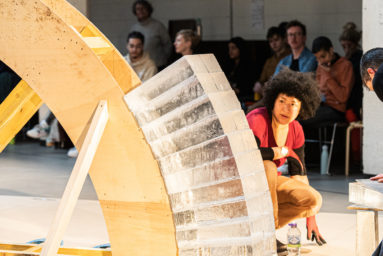 Blocks of ice are stacked against a section of wooden former. A person is crouched next to it studying it. Audience in the background watch on.
