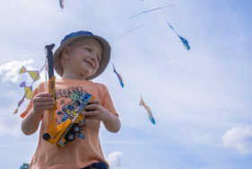 Outdoors. A smiling child holds a toy crane while the Fish swim above in the sky.