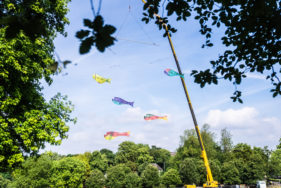 Fish Mobile, a giant kinetic sculpture, hanging in Rotherham park. A crane reaches high above the trees, about 70 metres in the air. Five fish hang from it suspended on horizontal bars. It's a bright and warm summer day.
