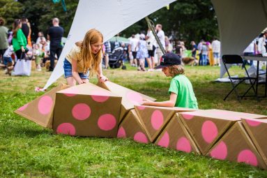 Outdoors, daytime on the grass. Two children play with a large pink spotty cardboard scultpure made of many prisms. They are working together to build something new.