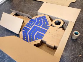 Indoors. A pile of unassembled Twist cardboard prism are stacked inside a tailored cardboard box. Rolls of tape and gloves are ready to pack too. A blue and yellow pattern is painted on the prisms.