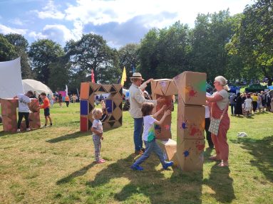 Outdoors. Daytime in a sunny park. Blue sky and trees line the background, with lots of people, stalls, flags and activity. In the foreground adults and children play with a long twist sculpture, trying to build a tall square for the children to jump through.
