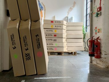 Indoors. A pile of long cardboard boxes sit on top of a pallet. On each box it says Kaleider's Let's Twist and there is a different colourful symbol on the side to indicate what is inside - a yellow pacman face, a green paint splat, a pink dot.