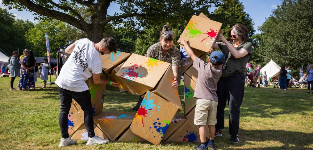 Outdoors, in a green park. Smiling people of different ages and sizes work together to build something with a large cardboard colourfully painted Twist sculpture.
