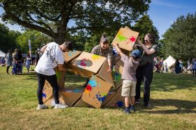 Outdoors, in a green park. Smiling people of different ages and sizes work together to build something with a large cardboard colourfully painted Twist sculpture.