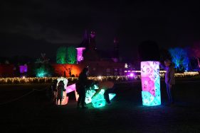Outdoors. Nightime. A family plays with glowing geometric sculptures in different shapes and colours. People are working together to move them. Glimpses of lit buildings and trees form a backdrop.