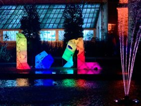 Outdoors. Nightime. A cluster of light up sculptures sit in front of glasshouse. The geometric shapes create vertical, horizontal, diagonal lines in the darkness. Rainbow coloured and bright, their reflection shimmers in the pond next to them.