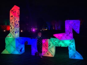 A 6 year old stands proudly in front of their glowing night-time creation. The cluster of shapes have been arranged together and on top on each other to build even taller and wider.
